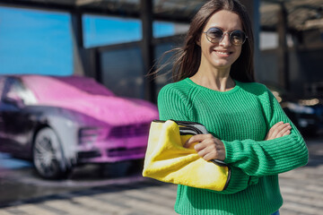 Portrait of young female in green sweater with yellow microfiber cloth and car that covered with...