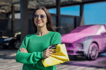Portrait of young female in green sweater with yellow microfiber cloth and car that covered with...