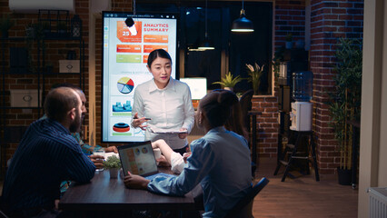 Asian company analyst explaining team revenue statistics in business meeting, financial report presentation, employee holding clipboard. Diverse coworkers discussing analytics data, talking