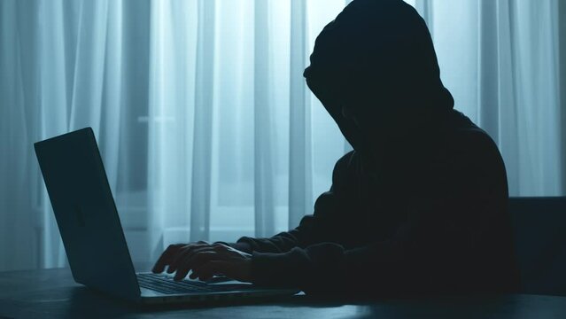 Silhouette of a computer hacker in a hood sitting at a laptop. Cyber attack.