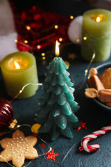 Burning Christmas tree candle with decor and treats on dark background, closeup