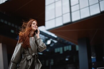 Fototapeta premium Woman walking around town and talking on the phone in trendy clothes against a backdrop of tall city buildings with a smile