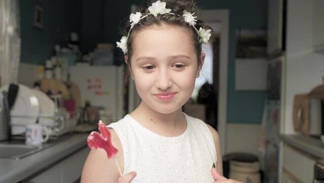Happy schoolgirl in white wreath of flowers. Beautiful Girl emotional face indoors close-up. Dream young girl at home. Funny kid licks candy on stick. Child smiles at camera. teenager eats lollipop