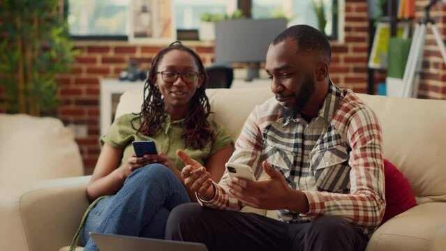 Young partners using mobile pohnes together on couch, browsing internet and scrolling through social media. People in love laughing at digital gadget, enjoying leisure activity relaxing.