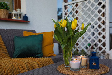 Soft pillows, blanket, burning candles and yellow tulips on rattan garden furniture outdoors
