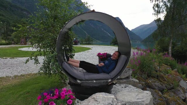 Cute girl smiling while relaxing inside circular pipe bed in spectacular landscape at Yrineset Olden glacial lake - Aerial flying through pipe before ascending to reveal majestic mountain landscape