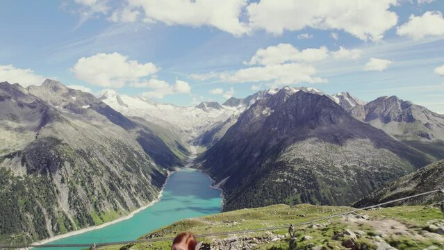Beautiful redhead girl standing on a wooden bridge waving. massive alpine mountain range scenery with glaciers in the distance. Big Schlegeis reservoir barrier lake in the valley on a sunny day 4K.