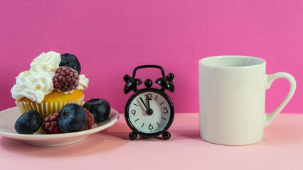 Obraz na płótnie Canvas Cupcake with cream and berries next to an alarm clock and a cup of black coffee. a clock and a cupcake on the table. A cup of coffee and a cupcake with an alarm clock on a pink table. Selective focus.