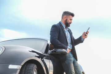 Handsome, bearded and attractive man consulting his telephone .leaning on his sports car