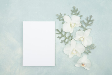 White orchids and blank stationery invitation on a light blue textured background flat lay