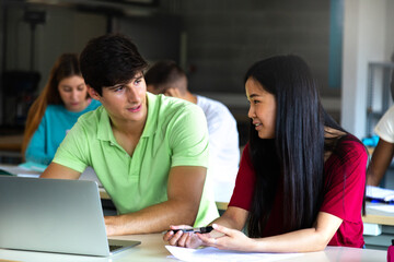 Caucasian teen boy and Asian teenage female high school students in class talking and working together using laptop.