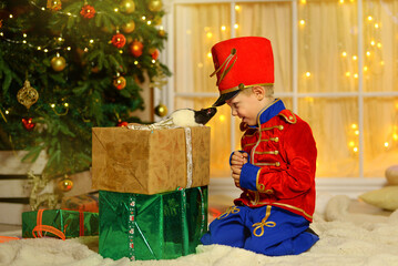 A boy in a nutcracker costume plays with the mouse symbol of the year. christmas tree and gifts