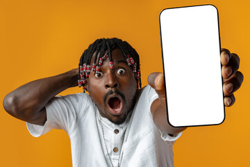 Excited young african man holding smartphone with white blank screen in hand on yellow studio background