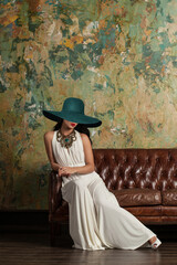 Beautiful young model in white evening gown and green wide broad brim hat sitting on luxury sofa