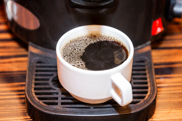 A white cup with prepared black coffee is in the coffee machine. Breakfasts and business meetings. Close-up.