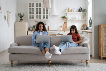 Modern introverted woman and teenage girl sits on sofa at home with laptops on laps. Unsociable Caucasian mother and child in casual clothes are addicted to gadgets and excessive use of internet