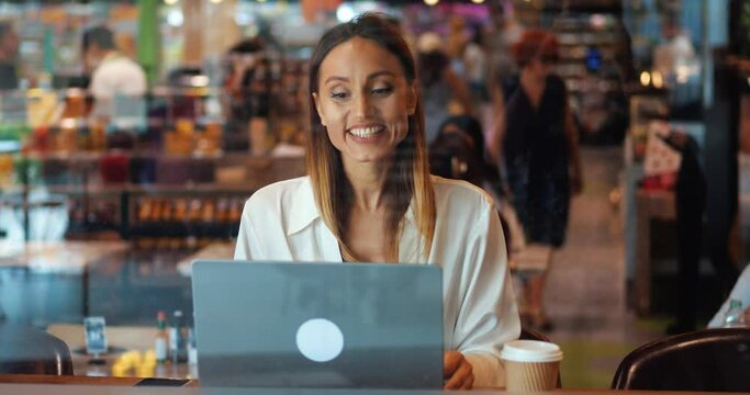 Charming multi-ethnic woman smiles a cheerful toothy smile, talking with her interlocutor via video link, sitting at cafe. Video conference. Online meeting with colleagues. Remote work. Telecommuting