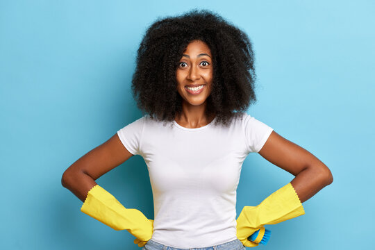 Indoor Shot Of Dark Skinned Housemaid With Exciting Smile, Wears Protected Gloves And White T-shirt, Poses Over Blue Background