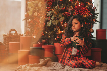 Portrait of candid asian smiling little girl in red plaid pajama sitting with presents at Xmas home
