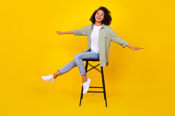 Fototapeta na wymiar Full size photo of shiny young girl wavy hair sit stool spread hands smile wear stylish khaki clothes isolated on yellow color background