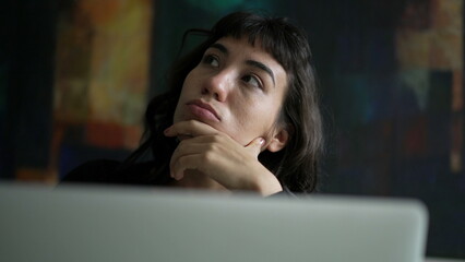 One pensive hispanic woman in front of laptop. A thoughtful latin person using computer thinking of an idea