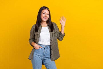 Portrait of good mood satisfied nice girl long hairdo wear khaki shirt hold arm in pocket waving hand isolated on yellow color background