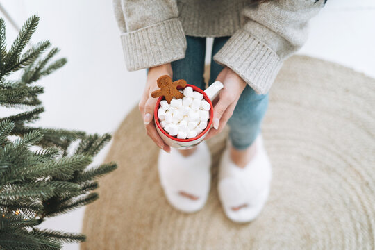 Young woman in cozy knitted sweater with cup of cocoa with marshmallow in room with Christmas tree at home