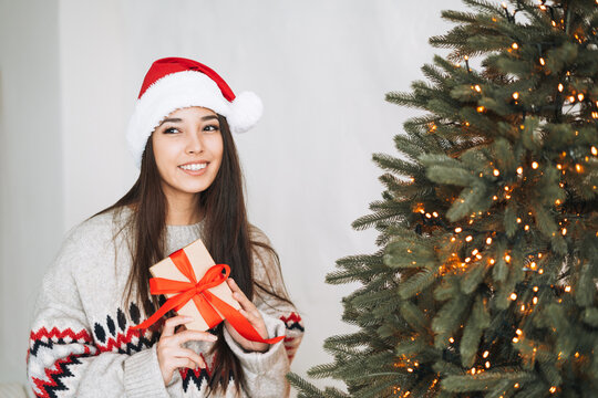 Young asian woman with dark long hair in Santa hat with present gift box with red ribbon in room with Christmas tree at home