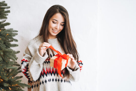 Young woman with dark long hair in cozy sweater with present gift box with red ribbon in room with Christmas tree at home