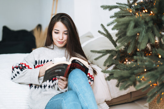 Young asian woman reading book on bed near Christmas tree at home