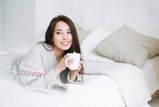 Young asian woman in cozy knitted sweater with cup of cocoa with marshmallow in bedroom at home