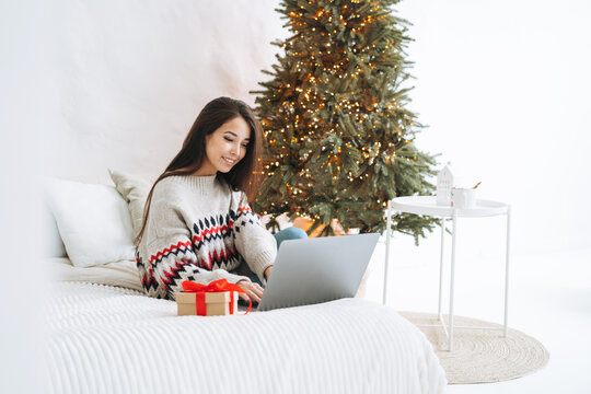 Young asian woman with dark long hair in cozy sweater using laptop on bed in room with Christmas tree at home