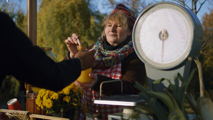 Obraz premium Elderly saleswoman stands at the stall, gives honey to taste. People pay for a jar of honey with cash. Local farmers market or autumn fair outdoors. Vegetarian and organic food. Agriculture.
