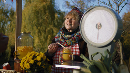 Elderly saleswoman stands at the stall, gives honey to taste. People pay for a jar of honey with...