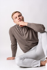 Caucasian blond model with a brown sweater on a white background, sitting on the floor with a seductive look