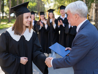 The teacher shakes hands with the student and presents the diploma outdoors. A group of university graduates.