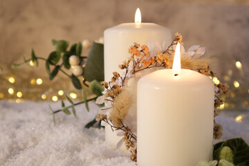 Two white candles decorated in Christmas decor on the snow against the background of lights