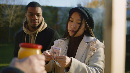 Diverse couple shopping at local farmers market. Asian woman pays for a jar of honey and puts it in eco bag. People walking on background. Autumn fair on weekend outdoors. Vegetarian and organic food.