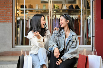 Two overjoyed Asian women enjoy talking while sitting on the stairs in front of the clothe store
