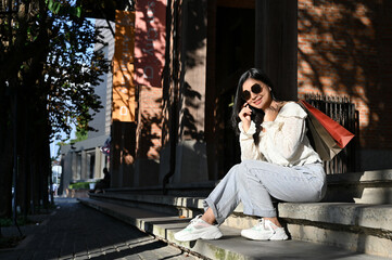 Attractive Asian female traveler with sunglasses and shopping bags sitting on street stairs.