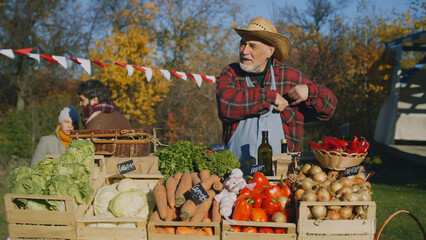 Senior farmer seller stands at the stall with fruits and vegetables, counts cash money. People shopping at local farmers market. Autumn fair. Organic food. Agriculture. Points of sale system.