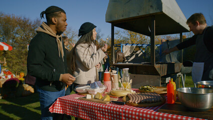 Multi ethnic couple at autumn fair or farmers markets. Asian woman and African American man trying cheese. Man cooks meat on grill. Holder of point of sale system. Outdoors picnic. Slow motion.
