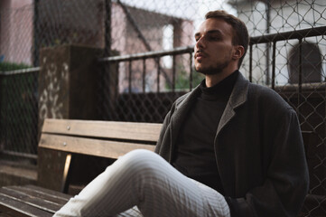 Obraz premium Thoughtful young man in dark coat sitting outdoors on wooden bench. Fashionable sad looking guy lonely on city street.