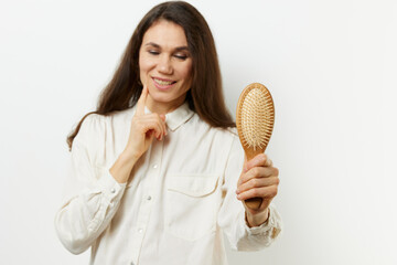 portrait of a beautiful, well-groomed, happy, adult woman on a light background in a white shirt, stretching her wooden massage comb into the camera, and looking at her very thoughtfully