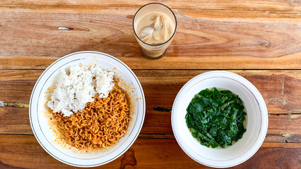 Bowl of moringa soup, instant fried noodles with rice, and iced coffee on a wooden table.