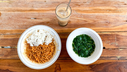 Bowl of instant fried noodles with rice, bowl of moringa soup and iced coffee on a wooden table.