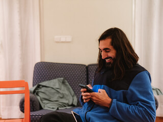 Young joyful caucasian man using mobile phone and smiling sitting at home on the couch