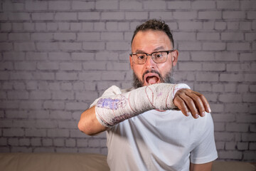Horizontal portrait, medium shot, of man with glasses and beard, white T-shirt and broken arm with plaster. With gesture of pain. He looks at his broken arm with expression of surprise, fear. Tenerife