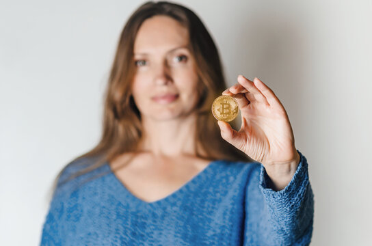 Joyful woman standing holding bitcoin paying attention to new digital cryptocurrency wearing blue sweater. Indoor studio shot isolated on white background.