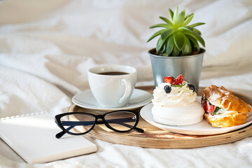 Cakes, coffee cup, notebook and eyeglass on wooden tray on bedding. Cozy breakfast in bed.